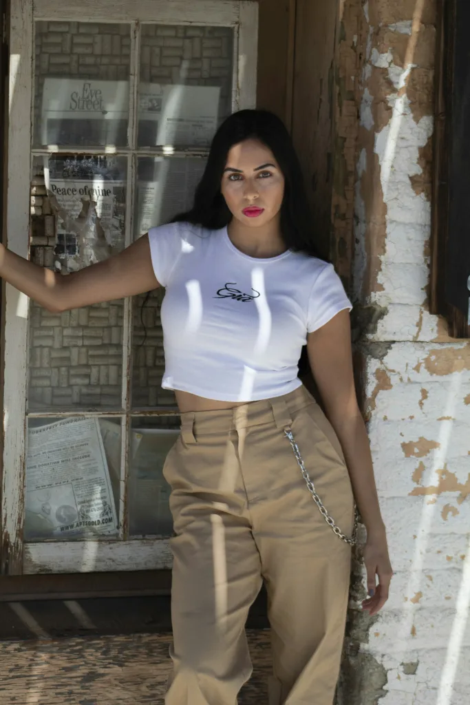 White cowgirl script baby tee with tan trousers, brick wall