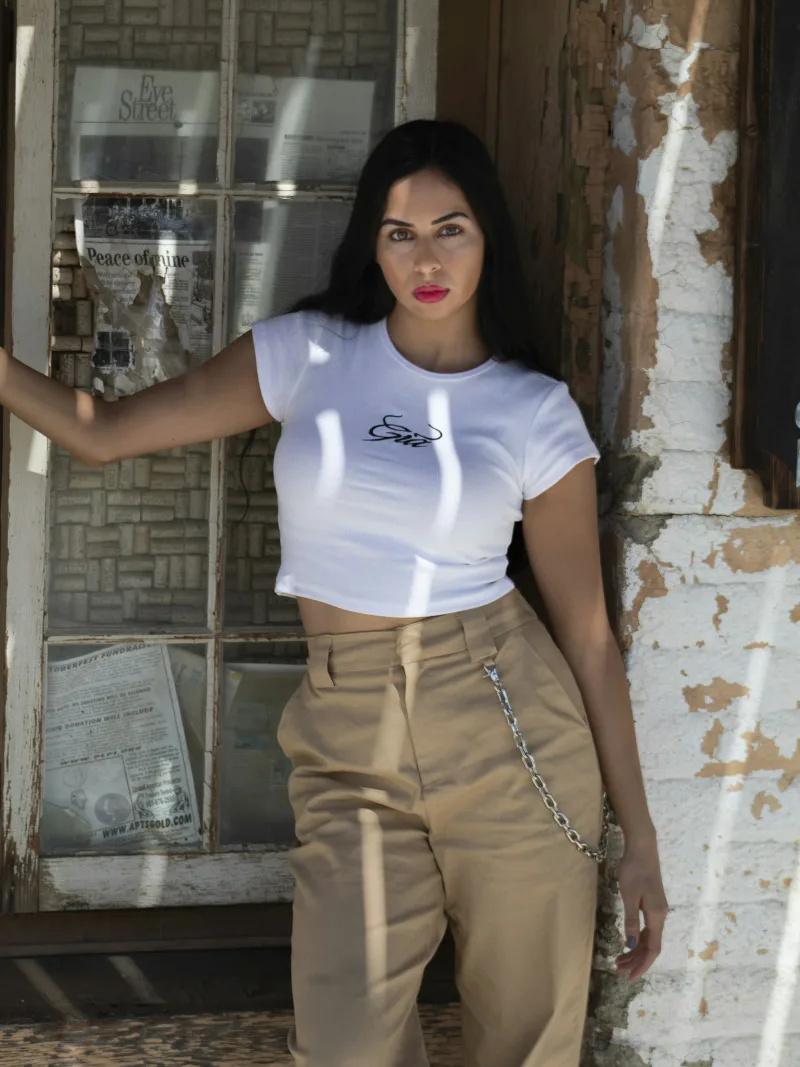 White cowgirl script baby tee with tan trousers, brick wall
